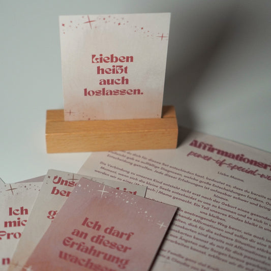 Ein Kartenhalter aus Holz präsentiert eine Karte mit der Aufschrift „Lieben heißt auch loslassen“. Daneben liegen auf einer weißen Unterlage das „Affirmationsritual – Besondere Affirmationskarten“ von Metra Fusion sowie eine ausgedruckte Liste mit Affirmationen, die die werdende Mutter unterstützen.