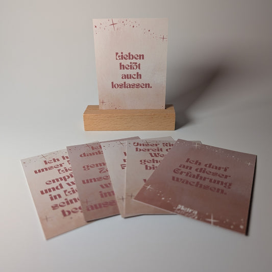Es sind fünf „Besondere Affirmationskarten“ von Metra Fusion zu sehen; eine Karte steht aufrecht in einem Holzhalter mit der Aufschrift „Lieben heißt auch loslassen“, während vier weitere Karten mit positiven Botschaften fächerförmig auf einer weißen Fläche ausgebreitet sind.